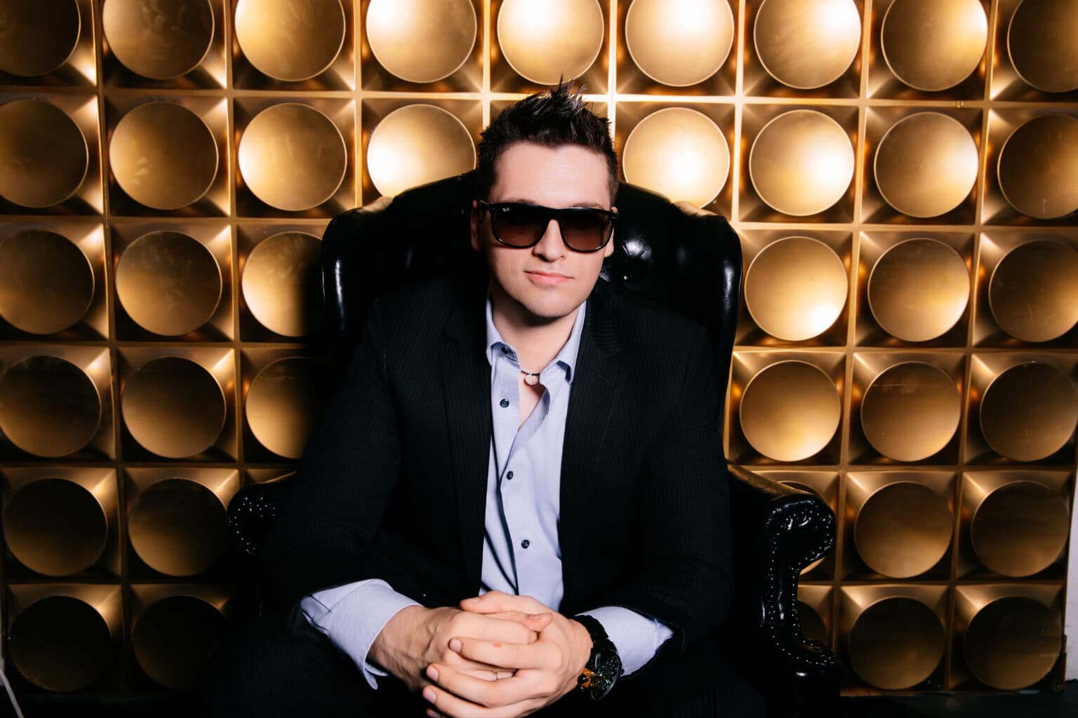 Stylish man with sunglasses in a black chair against golden geometric background for headshots.