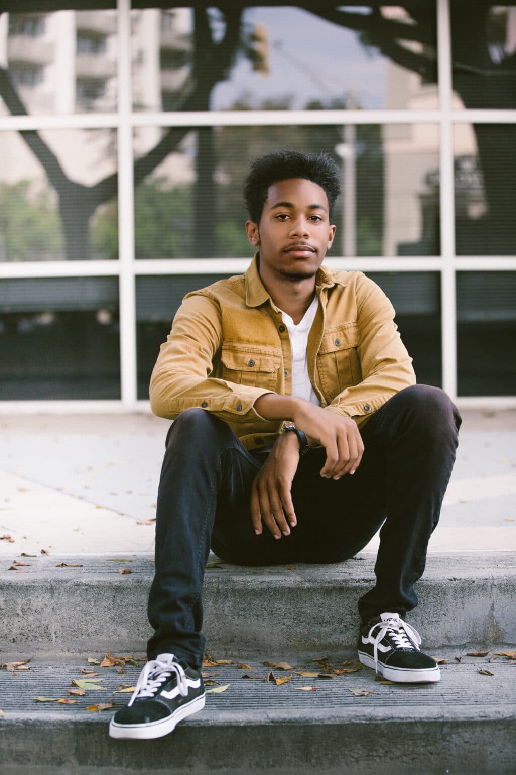 Young man sitting on curb outside modern building, casual attire, urban headshot for agency.