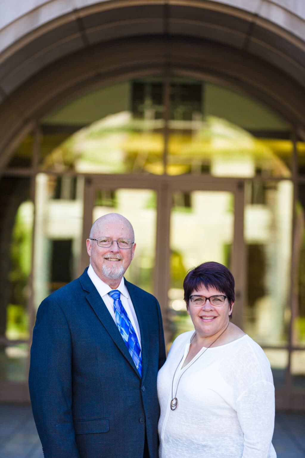Professional couple standing outdoors in front of modern building for corporate headshots.