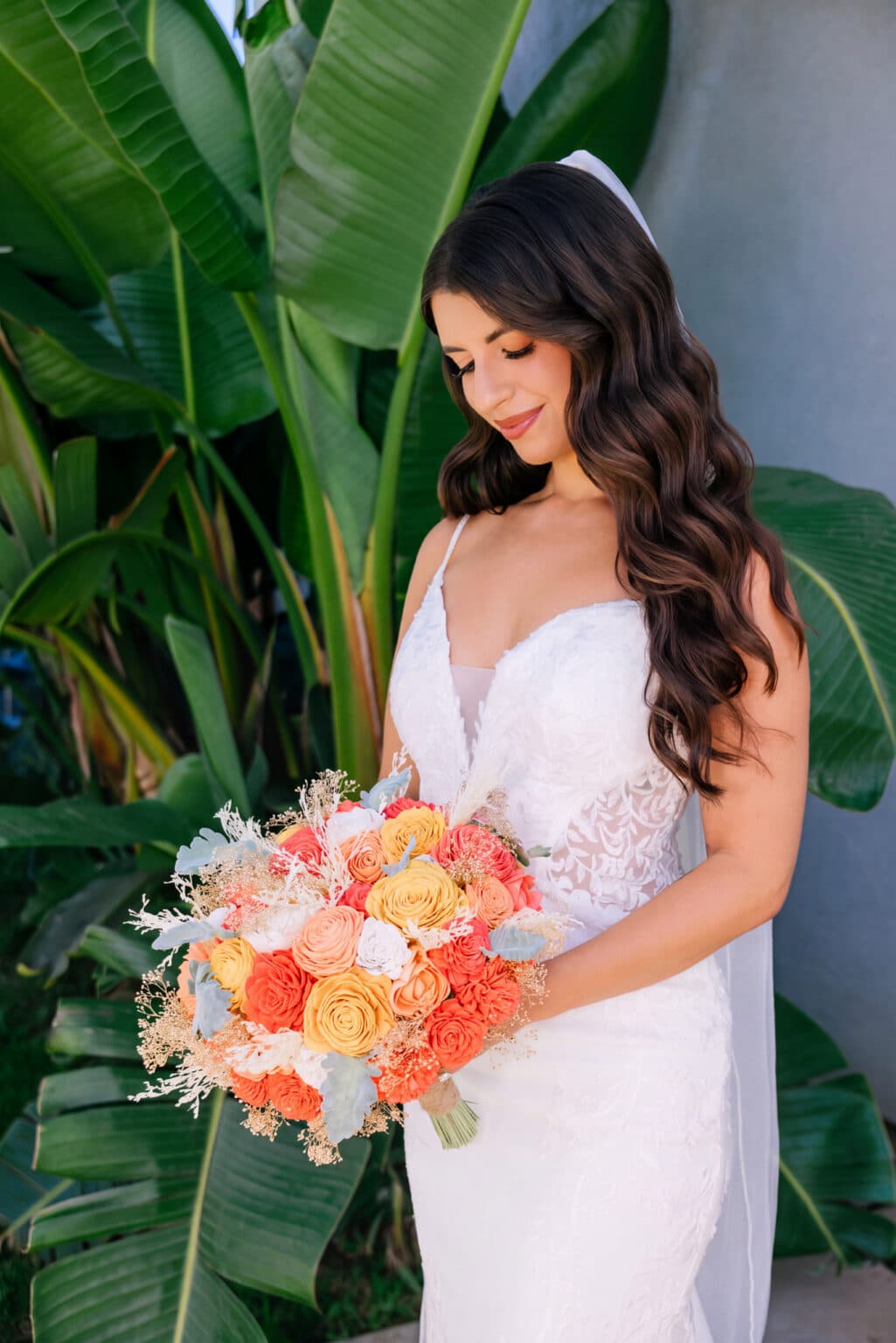 A bride in a white dress holds a bouquet of orange and yellow flowers, standing in front of large green leaves.