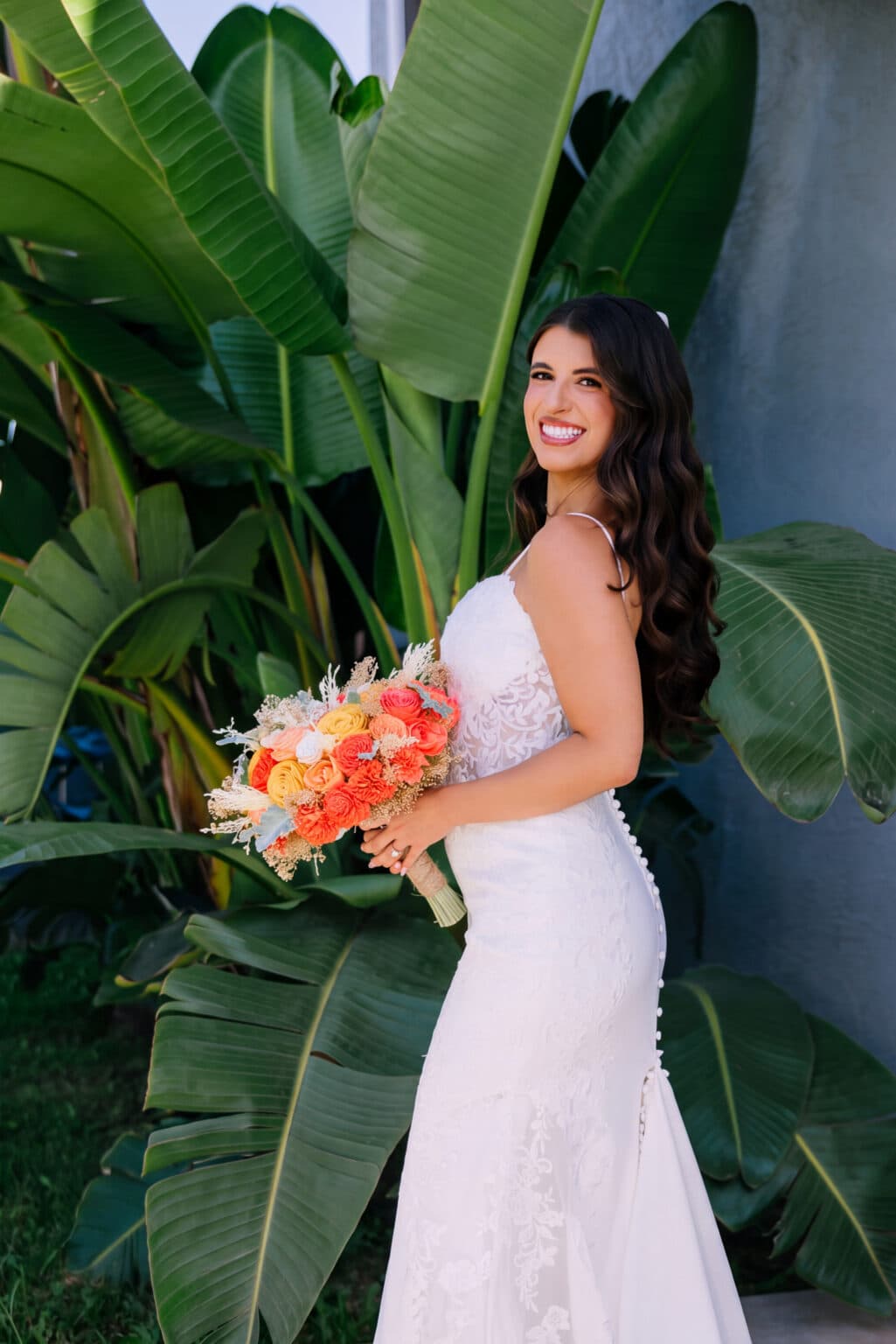 A woman in a white lace wedding dress holds a bouquet of orange and yellow flowers, standing in front of large green tropical plants.