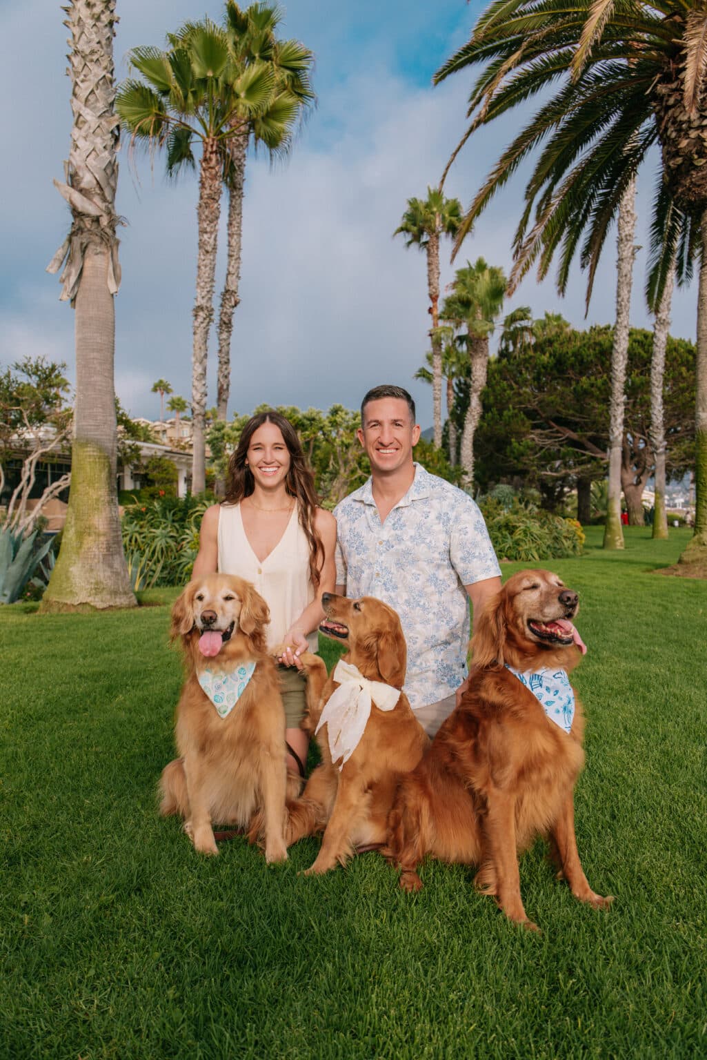 Outdoor family portrait of a couple smiling on a grassy lawn with their three Golden Retrievers wearing matching bandanas.